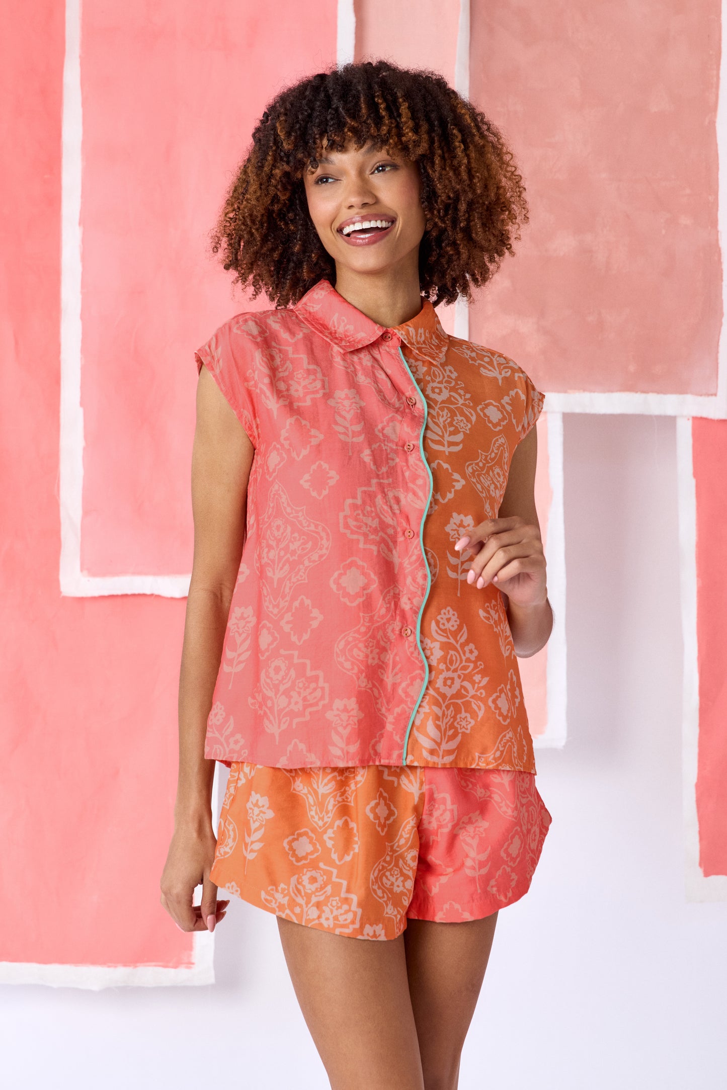 Model wearing color-blocked sleeveless blouse in coral and orange with a relaxed fit, featuring a white floral and geometric print, a pointed collar, button-front closure, and contrasting trim along the placket — CROSBY by Mollie Burch
