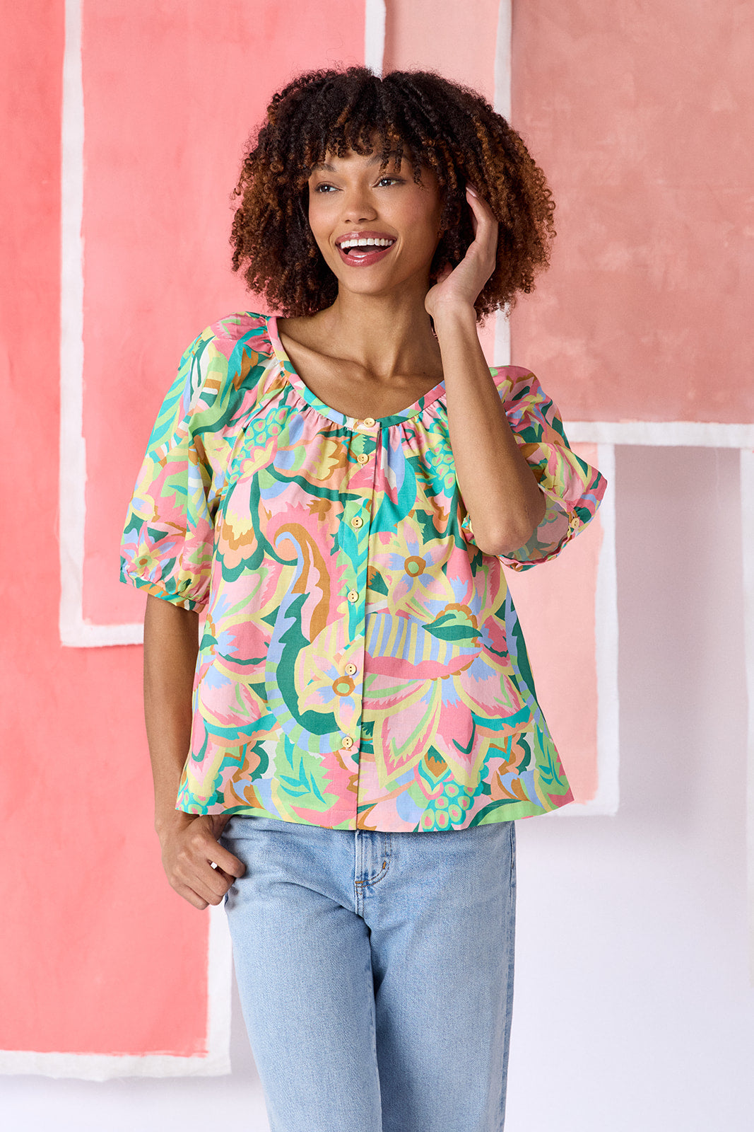 Model wearing a hip-length short sleeve top in a vibrant multicolor floral print, featuring a scoop neckline, button front, swingy silhouette, and short balloon sleeves — CROSBY by Mollie Burch