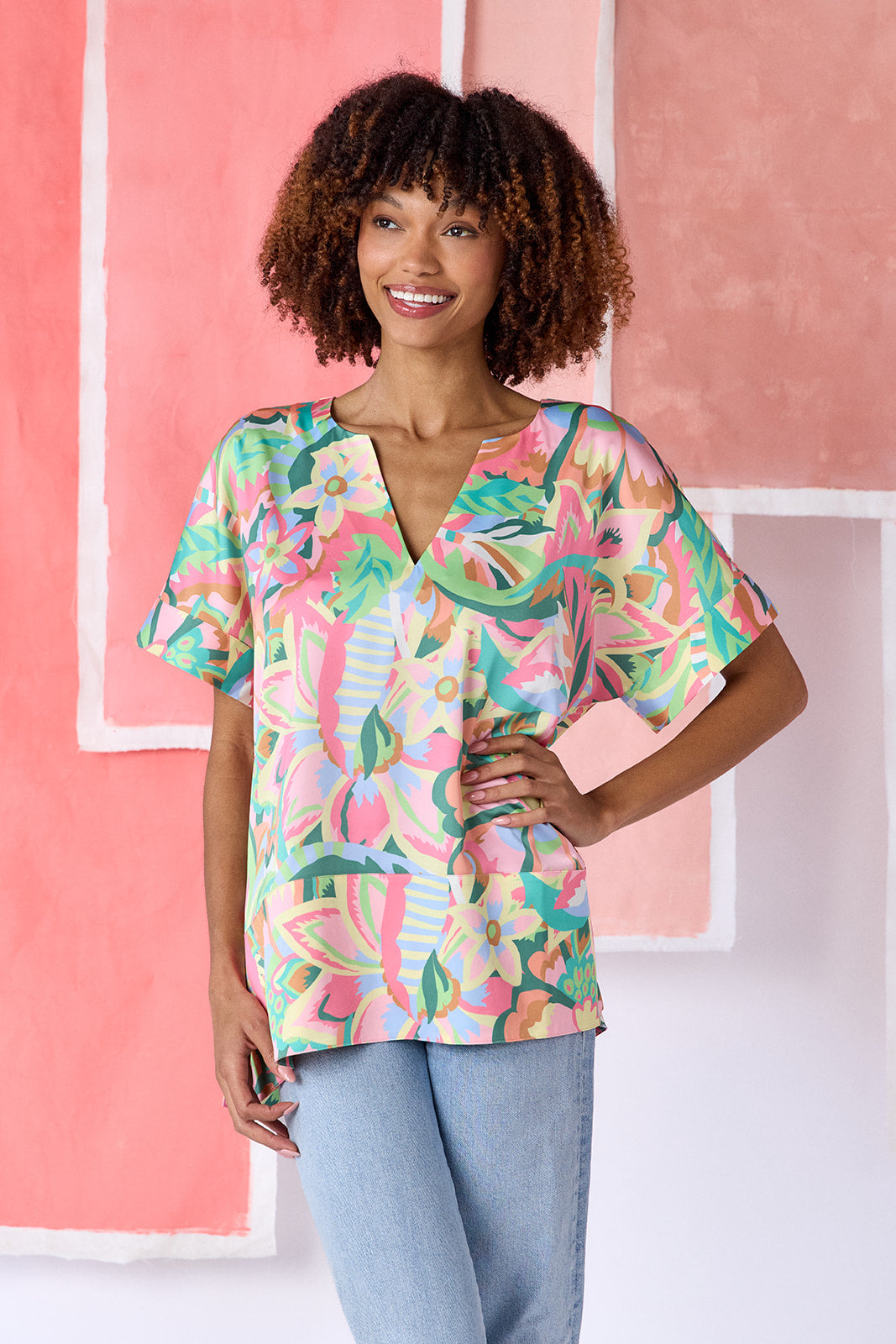 Model wearing top-of-the-thigh length tunic blouse in a vibrant multicolor floral print, featuring a breezy silhouette, split V-neck, and short dolman sleeves with seamed cuffs — CROSBY by Mollie Burch