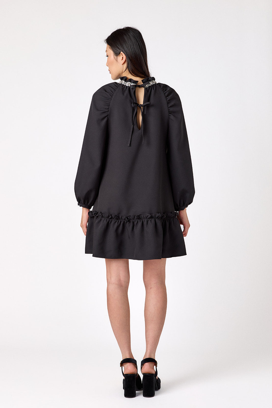 Flynn Dress in Black featuring rhinestone neckline, ruffle hem, and back tie detailing on model – CROSBY by Mollie Burch