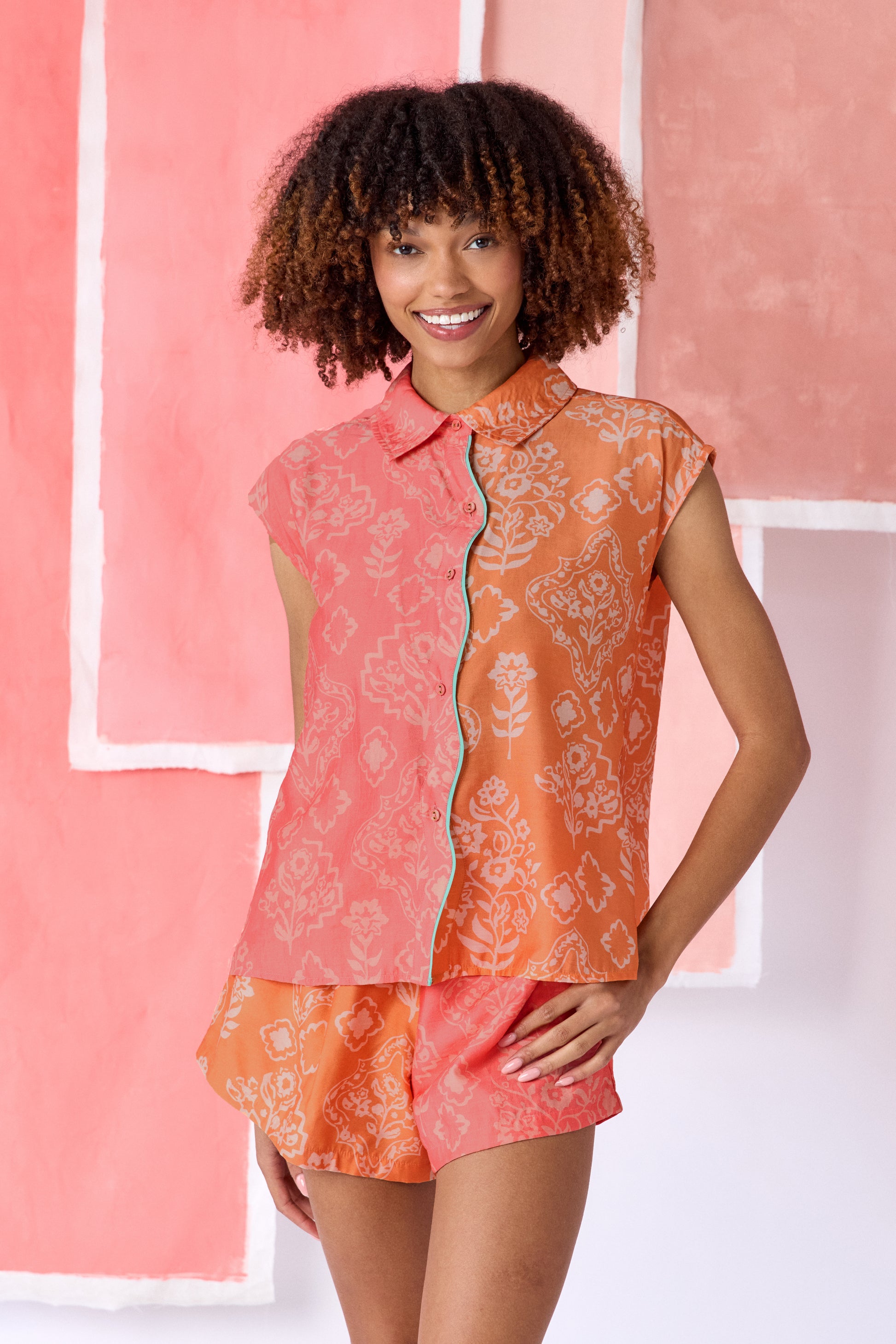 Model wearing color-blocked sleeveless blouse in coral and orange with a relaxed fit, featuring a white floral and geometric print, a pointed collar, button-front closure, and contrasting trim along the placket  — CROSBY by Mollie Burch