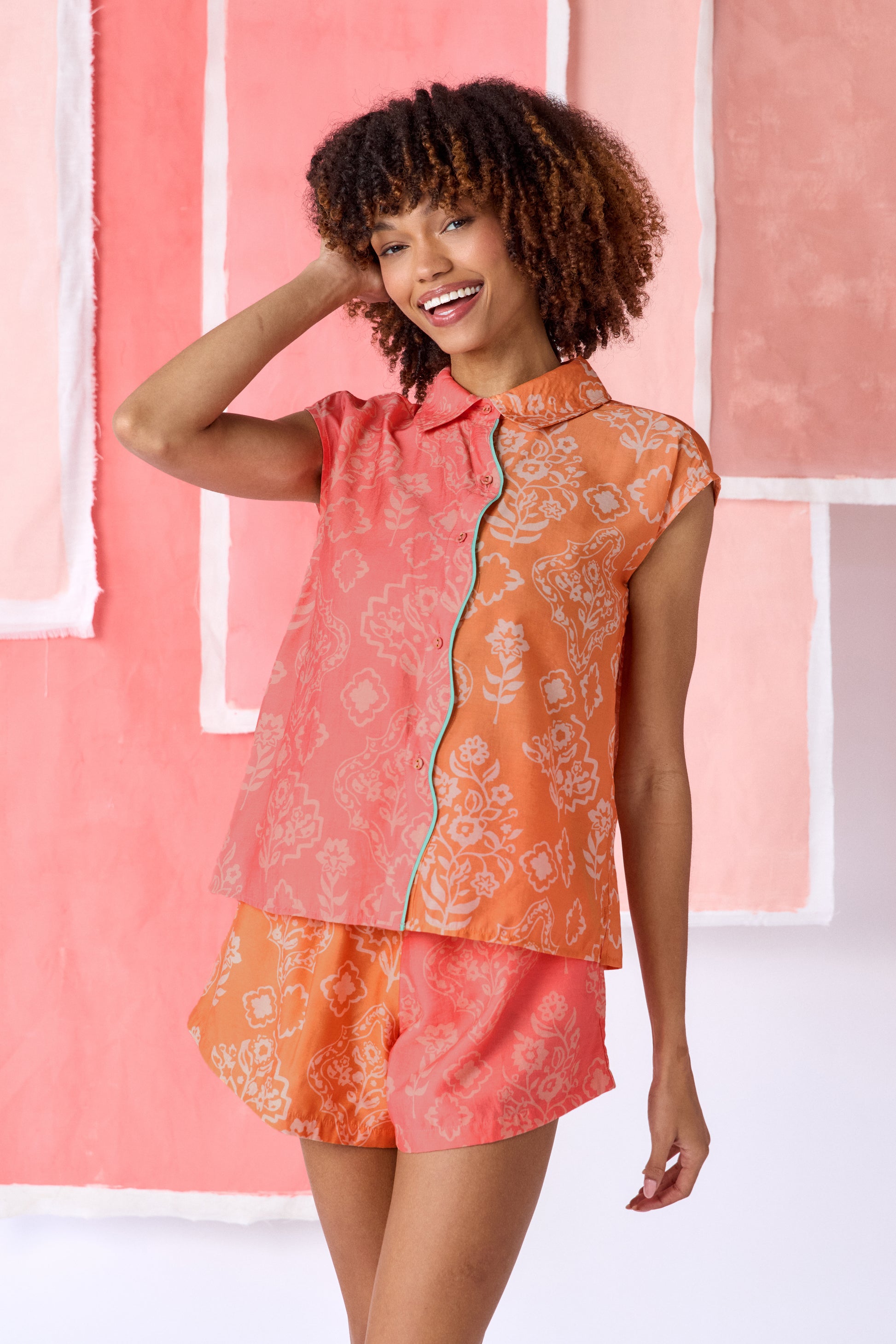 Model wearing color-blocked sleeveless blouse in coral and orange with a relaxed fit, featuring a white floral and geometric print, a pointed collar, button-front closure, and contrasting trim along the placket  — CROSBY by Mollie Burch