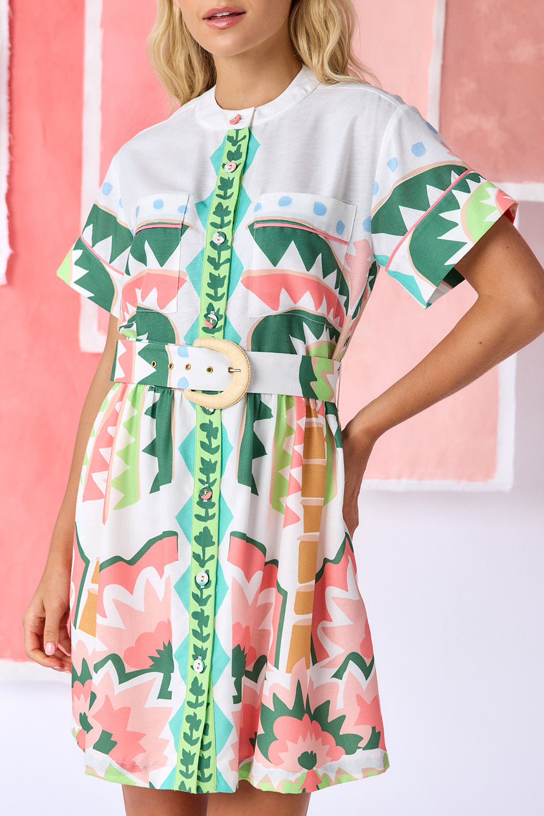 Model wearing above-the-knee, fit-and-flare dress with a full button front, short sleeves, and a relaxed fit. Features front breast pockets, contrast piping, and an optional belt with a natural raffia buckle. Finished in a colorful geometric print in green, coral, light blue, and tan tones — CROSBY by Mollie Burch