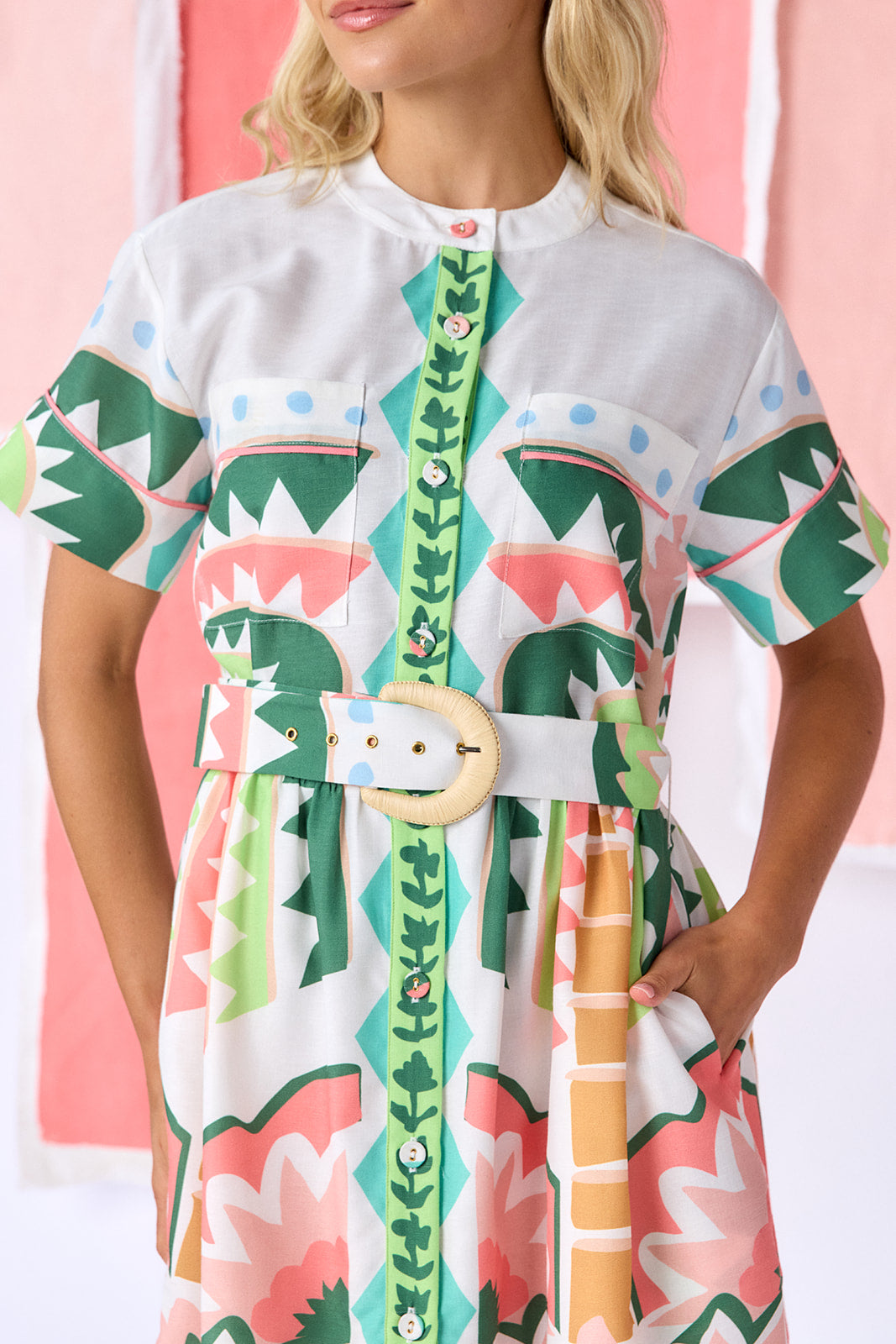 Model wearing above-the-knee, fit-and-flare dress with a full button front, short sleeves, and a relaxed fit. Features front breast pockets, contrast piping, and an optional belt with a natural raffia buckle. Finished in a colorful geometric print in green, coral, light blue, and tan tones — CROSBY by Mollie Burch