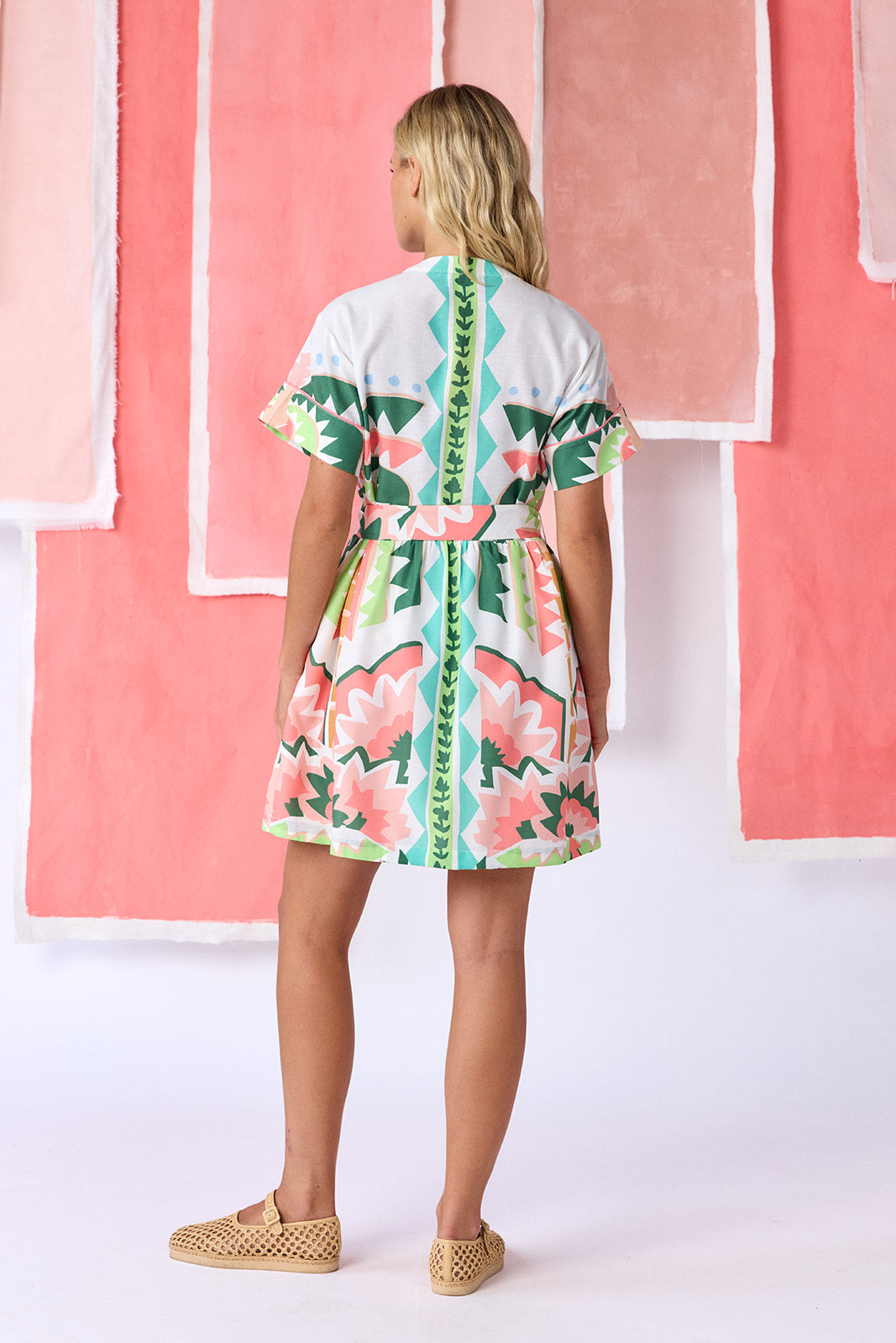 Model wearing above-the-knee, fit-and-flare dress with a full button front, short sleeves, and a relaxed fit. Features front breast pockets, contrast piping, and an optional belt with a natural raffia buckle. Finished in a colorful geometric print in green, coral, light blue, and tan tones — CROSBY by Mollie Burch