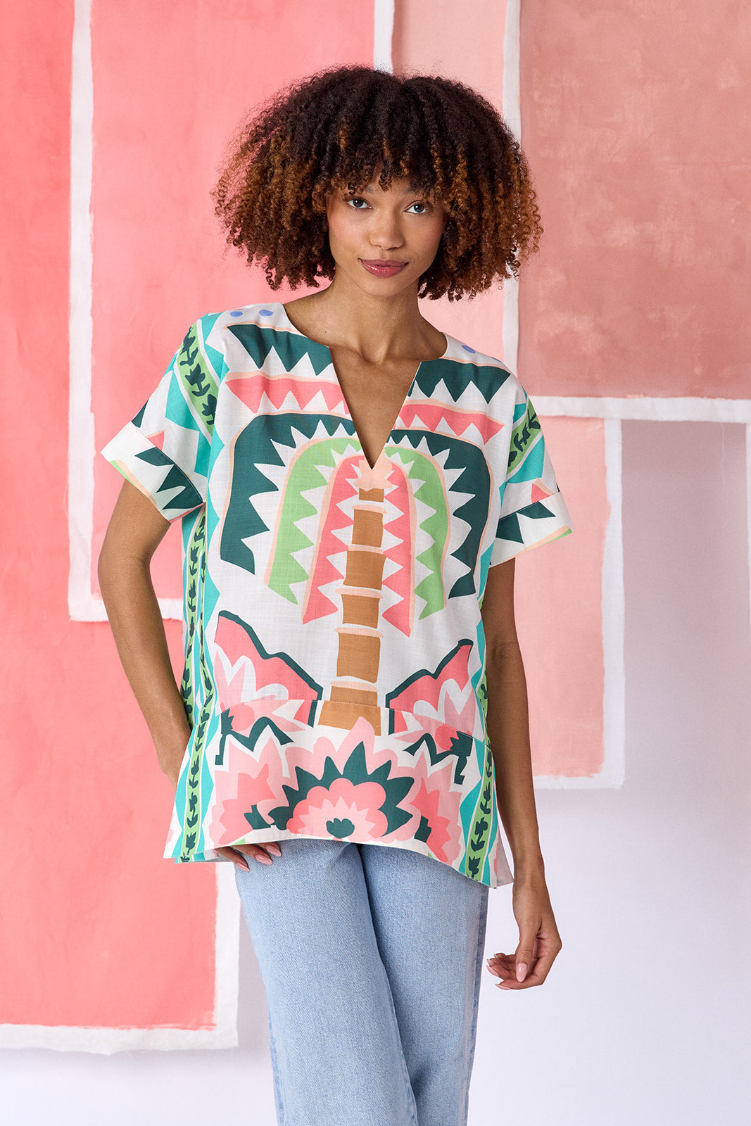 Model wearing top-of-the-thigh length short-sleeve tunic blouse with a breezy silhouette and split V-neck, in a colorful geometric print of green, coral, light blue, and tan tones — CROSBY by Mollie Burch