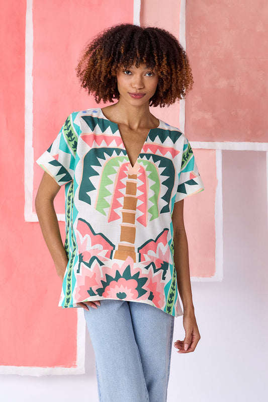 Model wearing top-of-the-thigh length short-sleeve tunic blouse with a breezy silhouette and split V-neck, in a colorful geometric print of green, coral, light blue, and tan tones — CROSBY by Mollie Burch