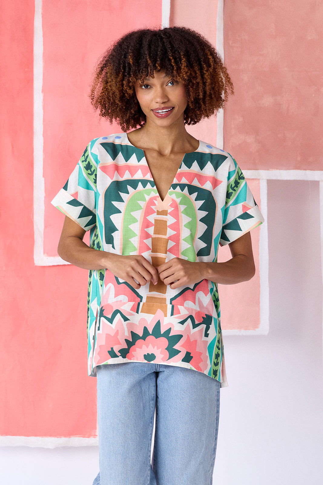 Model wearing top-of-the-thigh length short-sleeve tunic blouse with a breezy silhouette and split V-neck, in a colorful geometric print of green, coral, light blue, and tan tones — CROSBY by Mollie Burch