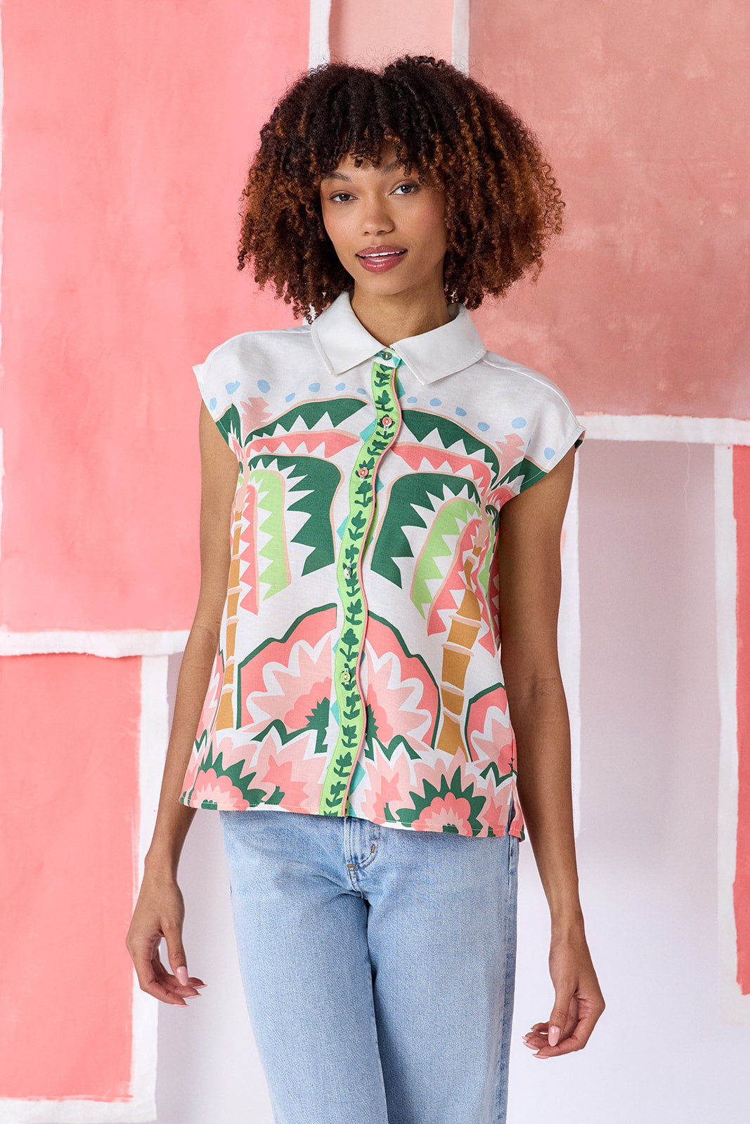 Model wearing a hip-length relaxed-fit top with cap sleeves, shirt collar, and a squiggle button placket with contrast trim, in a colorful geometric print of green, coral, light blue, and tan tones — CROSBY by Mollie Burch