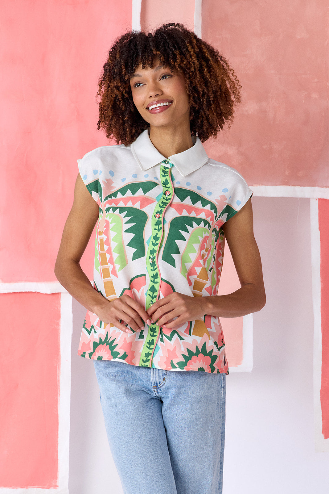 Model wearing a hip-length relaxed-fit top with cap sleeves, shirt collar, and a squiggle button placket with contrast trim, in a colorful geometric print of green, coral, light blue, and tan tones — CROSBY by Mollie Burch