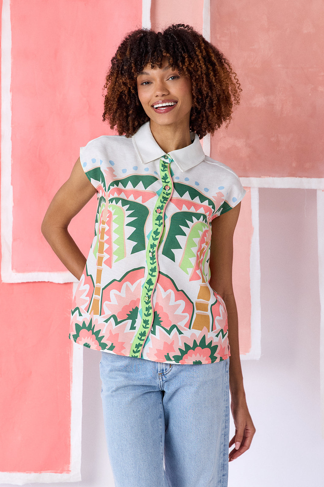 Model wearing a hip-length relaxed-fit top with cap sleeves, shirt collar, and a squiggle button placket with contrast trim, in a colorful geometric print of green, coral, light blue, and tan tones — CROSBY by Mollie Burch