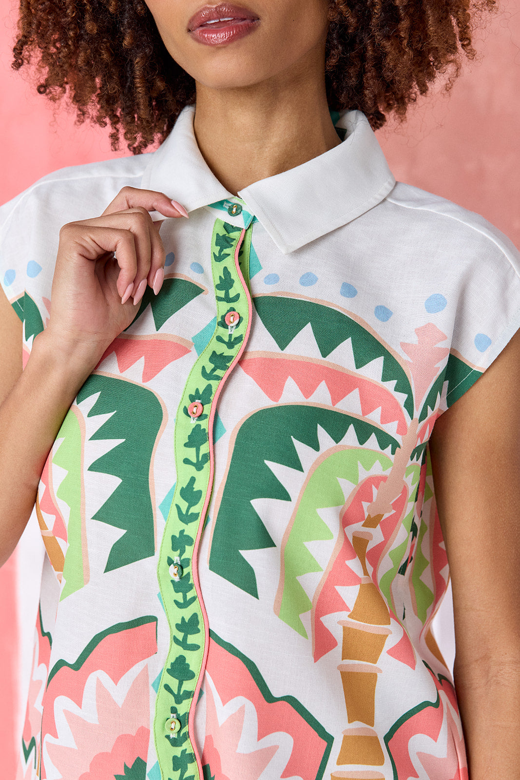 Model wearing a hip-length relaxed-fit top with cap sleeves, shirt collar, and a squiggle button placket with contrast trim, in a colorful geometric print of green, coral, light blue, and tan tones — CROSBY by Mollie Burch