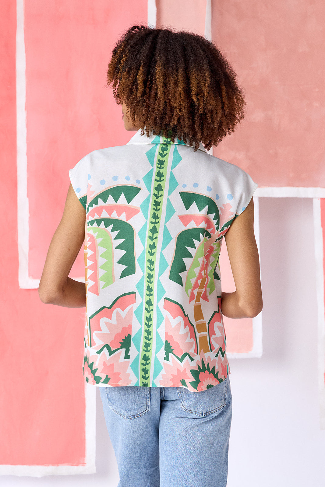 Model wearing a hip-length relaxed-fit top with cap sleeves, shirt collar, and a squiggle button placket with contrast trim, in a colorful geometric print of green, coral, light blue, and tan tones — CROSBY by Mollie Burch