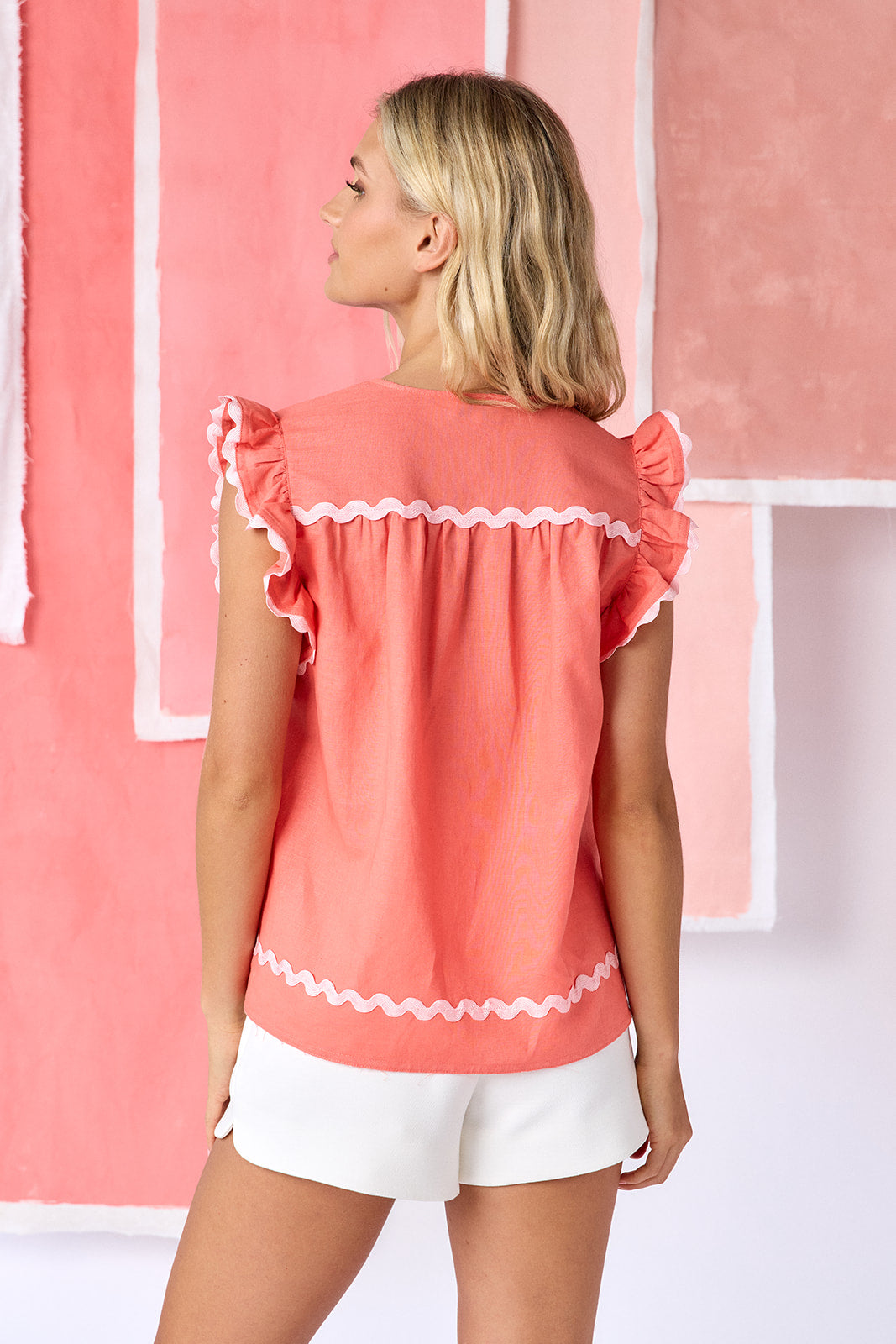 Model wearing sleeveless coral blouse with a relaxed, flowy fit, featuring ruffled cap sleeves with white trim, a ruffle-detail neckline, pintuck pleats at the chest, and a short button-front placket, finished with a white scalloped hem trim  — CROSBY by Mollie Burch