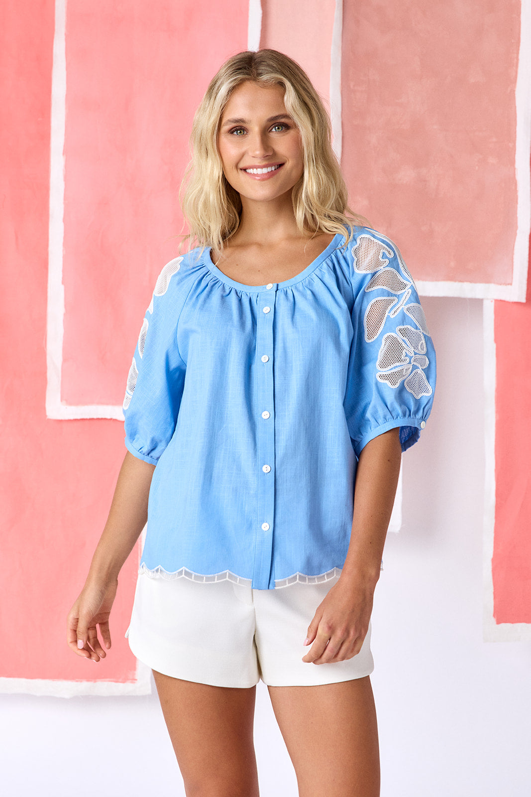 Model wearing light blue, hip-length top with a scoop neckline, button front, and a swingy silhouette. Features short balloon sleeves with sheer netting appliqué and a scallop-embroidered hem — CROSBY by Mollie Burch