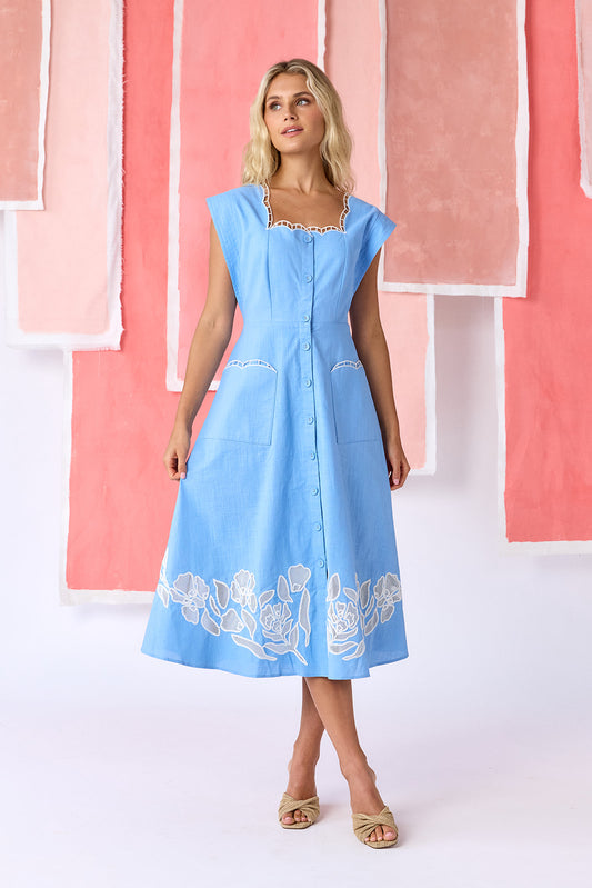 Model wearing a ight blue, mid-calf fit-and-flare dress with a square neckline, cap sleeves, and a full button front. Features a swingy skirt with sheer netting appliqué on the front and scallop-embroidered trim along the neckline and pocket openings — CROSBY by Mollie Burch