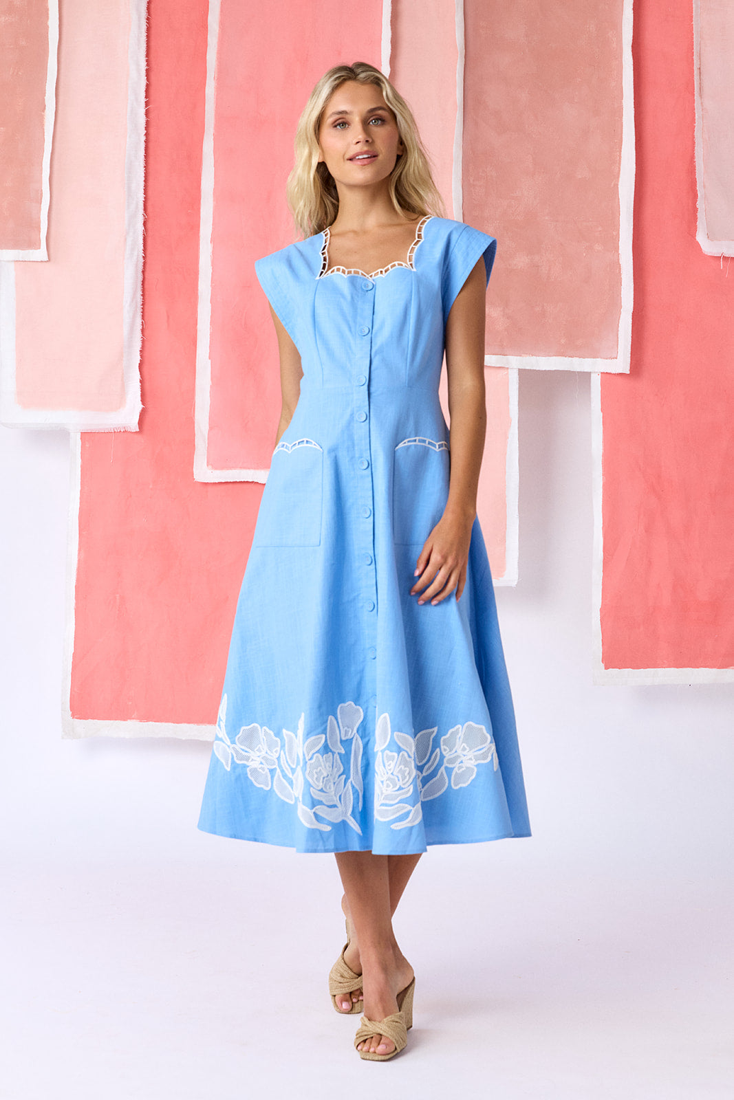 Model wearing a ight blue, mid-calf fit-and-flare dress with a square neckline, cap sleeves, and a full button front. Features a swingy skirt with sheer netting appliqué on the front and scallop-embroidered trim along the neckline and pocket openings — CROSBY by Mollie Burch