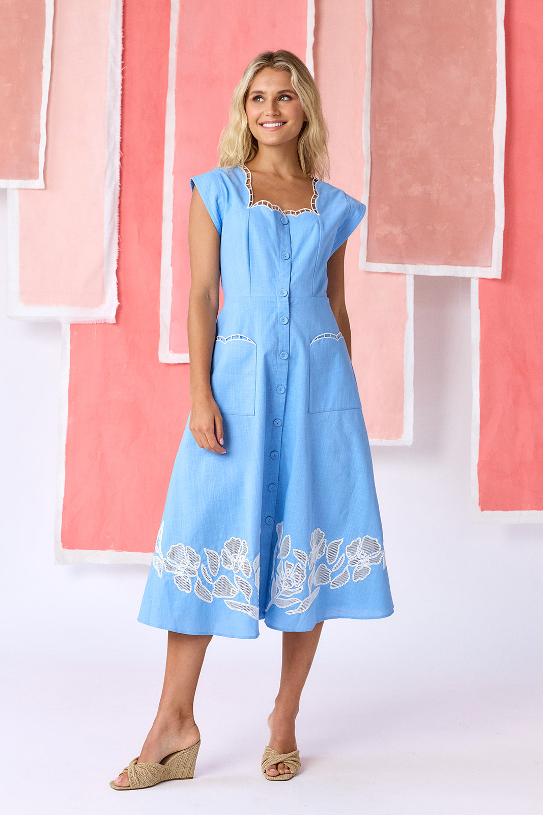 Model wearing a ight blue, mid-calf fit-and-flare dress with a square neckline, cap sleeves, and a full button front. Features a swingy skirt with sheer netting appliqué on the front and scallop-embroidered trim along the neckline and pocket openings — CROSBY by Mollie Burch