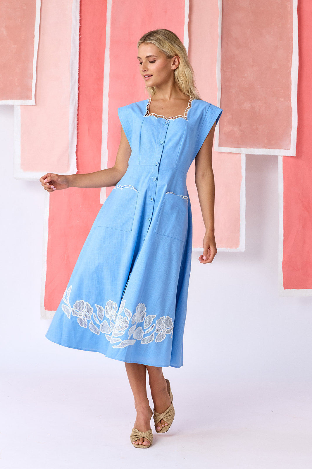 Model wearing a ight blue, mid-calf fit-and-flare dress with a square neckline, cap sleeves, and a full button front. Features a swingy skirt with sheer netting appliqué on the front and scallop-embroidered trim along the neckline and pocket openings — CROSBY by Mollie Burch