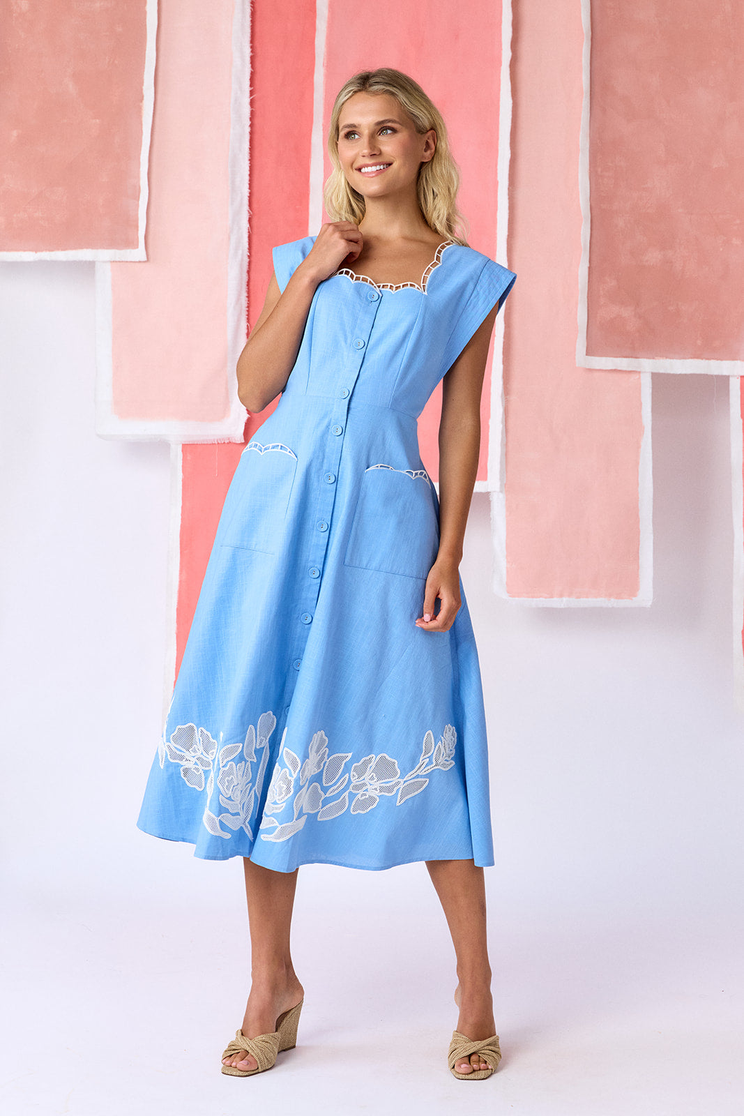 Model wearing a ight blue, mid-calf fit-and-flare dress with a square neckline, cap sleeves, and a full button front. Features a swingy skirt with sheer netting appliqué on the front and scallop-embroidered trim along the neckline and pocket openings — CROSBY by Mollie Burch